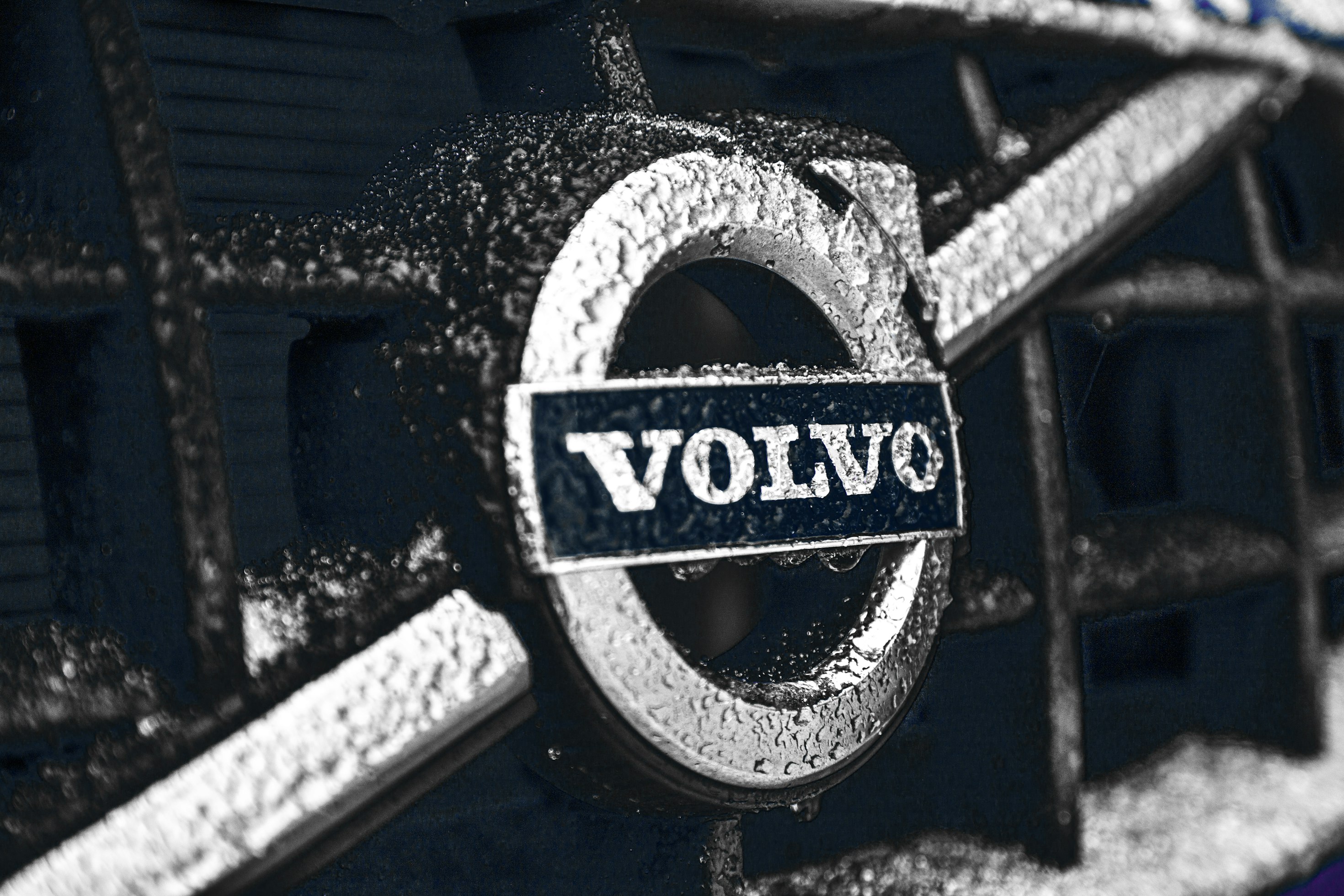 Close crop of a rain-beaded Volvo iron mark on a dark grille
